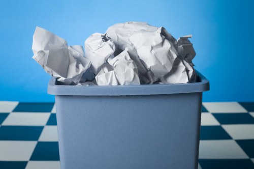 A close-up of a rectangular blue plastic bin filled with crumpled white and grey paper waste, placed on a floor with a black and white checkered pattern. The paper varies in size and texture, with some pieces showing creases and folds, indicating they are used or discarded documents. The background features a plain, solid blue wall, providing a simple and neutral backdrop that emphasizes the bin and its contents. This image represents typical rubbish collection or waste disposal scenes, relevant to professional rubbish removal services in Paddington. The scene appears to be indoors, possibly in a waste collection or storage area, with natural or soft lighting highlighting the textures of the paper and the smooth surface of the bin. As part of the context for Commercial Waste Removal Paddington, the image illustrates the type of waste material they handle, including paper waste from office or business premises, stored temporarily before disposal or recycling processes.