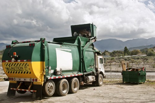 A large green waste collection truck with a black and yellow caution strip on the rear, parked on a gravel surface against a backdrop of distant mountains and cloudy sky. The truck features a hydraulic lifting mechanism for rubbish compaction, and a small green skip filled with mixed waste materials is positioned nearby on the right side. The scene is outdoors, likely in a waste management or disposal yard, illustrating a typical rubbish removal operation that a service such as Commercial Waste Removal Paddington might perform in the area around Westminster or Paddington, London. The environment appears overcast with diffuse lighting, highlighting the industrial equipment and empty open space suitable for rubbish collection and disposal activities.