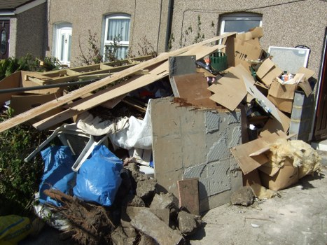A large pile of mixed household waste and construction debris is piled against a residential building's exterior wall, positioned on a paved pavement. The heap includes broken drywall sheets, concrete blocks, cardboard boxes, and wood planks, some of which are leaning or stacked imperfectly. There are also plastic bags and loose materials among the clutter, with the debris varying in colour from grey concrete and brown cardboard to natural wood tones and scattered white coverings. The background shows the lower parts of two residential windows with white frames, and the building's exterior is finished in a beige textured surface. The overall scene indicates a typical rubbish removal situation in a residential area, with the waste likely awaiting collection or disposal through a professional service such as Commercial Waste Removal Paddington, operating near Paddington in London.