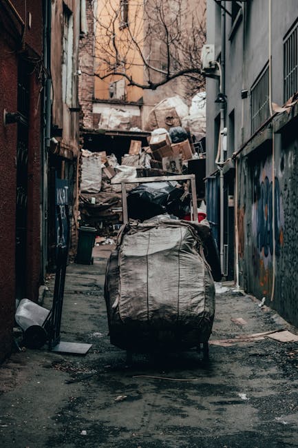 A collection of overflowing waste bins and scattered rubbish is positioned on a paved sidewalk in front of a commercial building, illustrating a scene of illegal or unmanaged waste accumulation. The c